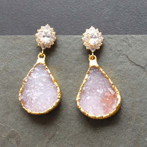 May include: A pair of gold-toned drop earrings. Each earring features a teardrop-shaped, light purple druzy stone with a gold-colored trim. The top of each earring has a cluster of small, clear crystals surrounding a larger, clear crystal.