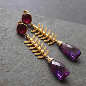 May include: A pair of gold-colored earrings with a fishbone design. Each earring features a square, red gemstone at the top, a gold fishbone-shaped chain, and a faceted, teardrop-shaped purple gemstone at the bottom. The earrings are displayed on a dark gray surface.