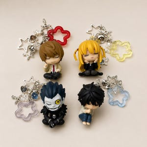 May include: Four anime character keychains featuring chibi-style figures of Light Yagami, Misa Amane, Ryuk, and L. Each figure is attached to a silver-tone metal chain with various charms, including clear plastic stars in red, yellow, blue, and purple.