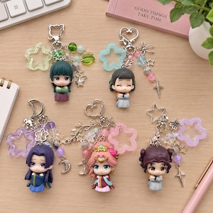 May include: Five character keychains featuring chibi-style figures with diverse hair colours and traditional-style clothing. Each keychain includes a metal clasp, a translucent star-shaped charm, and various small beads, stars, and moon-shaped metal accents.