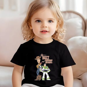 Puede incluir: Una camiseta negra con un gr&aacute;fico de Buzz Lightyear y Woody de Toy Story. El texto "Buzz Woody" est&aacute; impreso en marr&oacute;n en la camiseta.