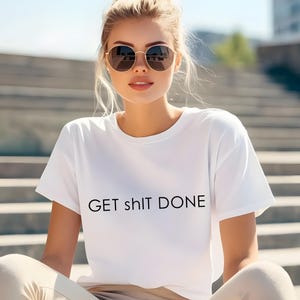 May include: White t-shirt with the text "GET sh!t DONE" in black. The shirt is short-sleeved and appears to be made of a soft material. The person in the image is wearing sunglasses and sitting outside.
