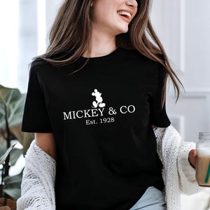 May include: Black t-shirt with the text "MICKEY & CO Est. 1928" in white. The design includes a silhouette of Mickey Mouse. The shirt is worn by a person holding a glass of beverage with a green straw.