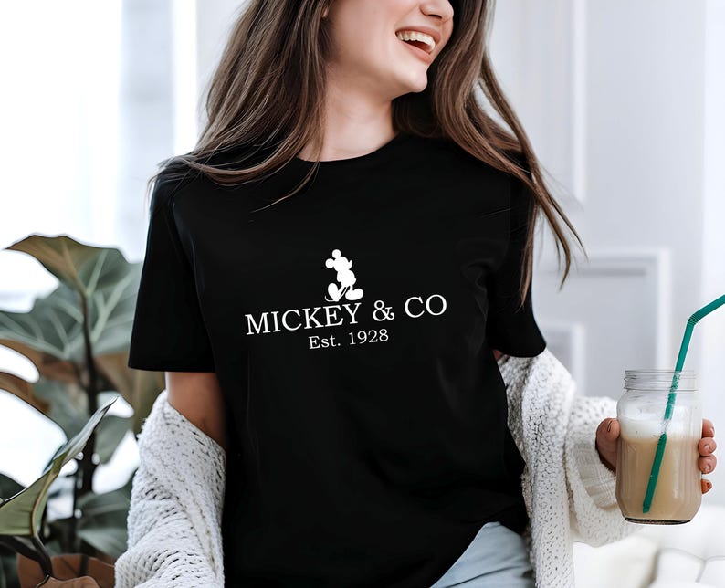 May include: Black t-shirt with the text "MICKEY & CO Est. 1928" in white. The design includes a silhouette of Mickey Mouse. The shirt is worn by a person holding a glass of beverage with a green straw.