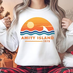 May include: A white long-sleeved sweatshirt featuring a graphic of a sunset over ocean waves with a shark fin. The text AMITY ISLAND 1975 is printed in orange below the waves. The design uses shades of yellow, orange, and blue on the white fabric.