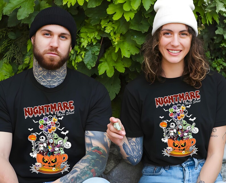 May include: Black t-shirts with a Halloween-themed graphic. The design features the text "Nightmare on Main Street" above a pumpkin-shaped teacup filled with cartoon characters and balloons. Bats and other spooky elements complete the design.
