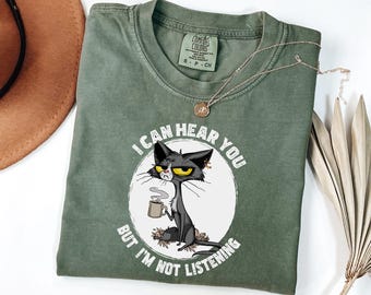 May include: Olive green t-shirt with a graphic of a grumpy black cat holding a mug, with the text "I CAN HEAR YOU BUT I'M NOT LISTENING". A gold necklace and a dried palm leaf are also visible.