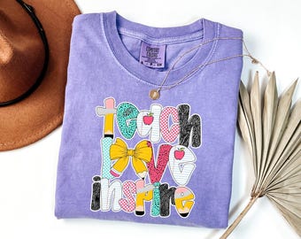 Puede incluir: Una camiseta lila Comfort Colors con las palabras "Teach Love Inspire" en un diseño colorido y juguetón. El diseño incluye un lazo amarillo, lápices y manzanas. También se ven un collar dorado y un sombrero marrón.