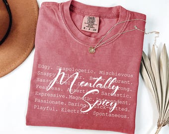 Puede incluir: Una camiseta de color rojo con la frase "Mentally Spicy" en escritura blanca. La camiseta también enumera varios adjetivos. Un collar plateado con un colgante está sobre la camiseta doblada.