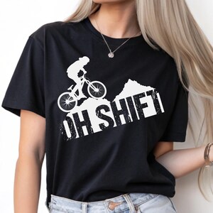 May include: A black short-sleeved t-shirt featuring a white graphic of a person riding a mountain bike over a hill. Below the graphic, the text "OH SHIFT" is printed in a bold, distressed white font. The shirt has a crew neckline.
