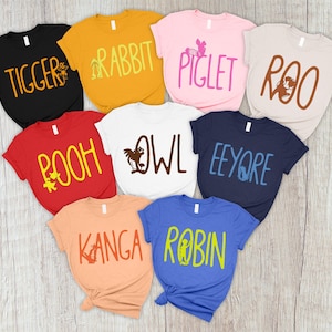 Può includere: Una collezione di magliette colorate con i nomi dei personaggi di una storia per bambini. Le magliette sono in vari colori, tra cui nero, giallo, rosa, beige, rosso, bianco, arancione e blu. Ogni maglietta ha un nome diverso stampato.