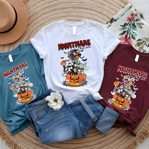 May include: Three t-shirts in teal, white, and burgundy, each featuring a Halloween-themed graphic. The design includes a jack-o'-lantern, characters, and the text "NIGHTMARE Main Street". The shirts are displayed with blue jeans and a straw hat.