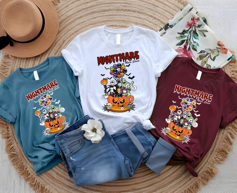 May include: Three t-shirts in teal, white, and burgundy, each featuring a Halloween-themed graphic. The design includes a jack-o'-lantern, characters, and the text "NIGHTMARE Main Street". The shirts are displayed with blue jeans and a straw hat.