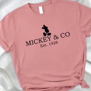 May include: A dusty rose-colored t-shirt featuring the text "MICKEY & CO Est. 1928" in black, with a silhouette of Mickey Mouse above the text. The shirt has rolled-up sleeves and a knotted hem.