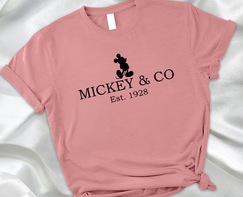 May include: A dusty rose-colored t-shirt featuring the text "MICKEY & CO Est. 1928" in black, with a silhouette of Mickey Mouse above the text. The shirt has rolled-up sleeves and a knotted hem.