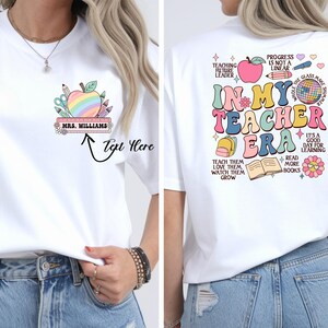 Può includere: Una t-shirt bianca con una grafica frontale di una mela arcobaleno con materiale scolastico e il testo Mrs. Williams. La grafica sul retro mostra il testo In My Teacher Era circondato da illustrazioni colorate di una mela, zaino e libro.