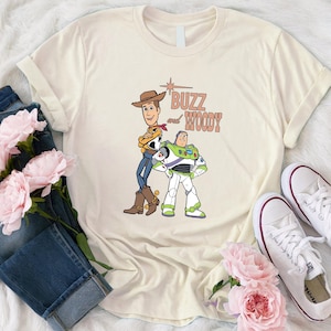Puede incluir: Una camiseta beige con un gr&aacute;fico de Woody y Buzz Lightyear de Toy Story. El texto "Buzz and Woody" est&aacute; impreso en marr&oacute;n en la camiseta.