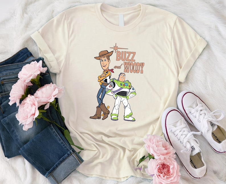 Puede incluir: Una camiseta beige con un gr&aacute;fico de Woody y Buzz Lightyear de Toy Story. El texto "Buzz and Woody" est&aacute; impreso en marr&oacute;n en la camiseta.