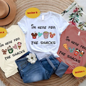 May include: Three t-shirts with the phrase "I'm here for the snacks" and various food-themed graphics. The shirts are white, cream, and coral. Blue jeans and a straw hat are also in the image.