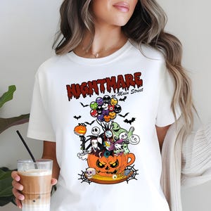 May include: White t-shirt featuring a colorful graphic design with the text "NIGHTMARE on Main Street." The design includes characters from The Nightmare Before Christmas, a pumpkin, bats, and balloons.