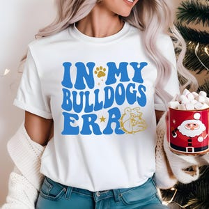 Può includere: Una t-shirt bianca a maniche corte con la scritta blu "IN MY BULLDOGS ERA", un'impronta di zampa dorata, una stella dorata e l'illustrazione di un bulldog dorato. È abbinata a una tazza rossa decorata con Babbo Natale e piena di marshmallow.