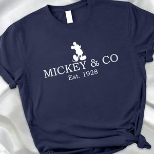 May include: Navy blue t-shirt featuring a white Mickey Mouse silhouette and the text "MICKEY & CO Est. 1928". The shirt is made of a soft material and has a classic crew neck design.