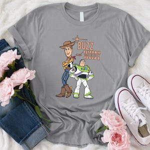 Puede incluir: Camiseta gris con un gr&aacute;fico de Buzz Lightyear y Woody de Toy Story. El texto "Buzz and Woody" est&aacute; impreso en naranja en la camiseta.