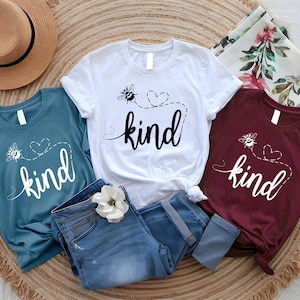 May include: Three t-shirts in teal, white, and burgundy, each with a bee graphic and the word "kind" in a script font. The shirts are paired with blue jeans and a straw hat, suggesting a casual, everyday style.