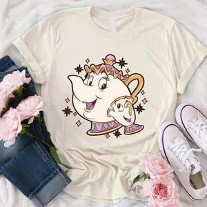 May include: Cream-colored t-shirt featuring a cartoon illustration of Mrs. Potts and Chip from Beauty and the Beast. The design includes a smiling teapot and teacup with decorative stars. The shirt is paired with blue jeans and white sneakers.