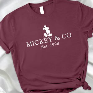 May include: A maroon t-shirt with rolled-up sleeves. The white graphic on the front features Mickey Mouse and the text "MICKEY & CO Est. 1928". The shirt is knotted at the bottom.