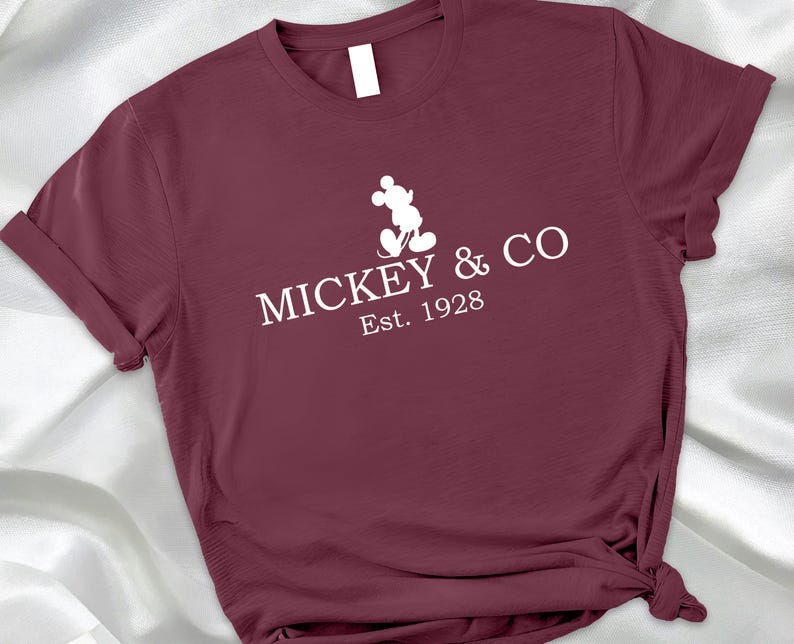 May include: A maroon t-shirt with rolled-up sleeves. The white graphic on the front features Mickey Mouse and the text "MICKEY & CO Est. 1928". The shirt is knotted at the bottom.