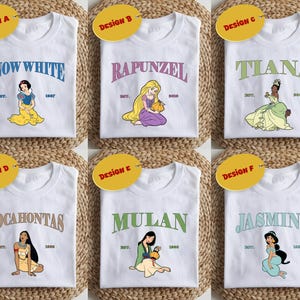 May include: Six white t-shirts with different Disney princess designs. Each shirt features a princess's name in a colourful banner and an illustration of the princess. The princesses featured are Snow White, Rapunzel, Tiana, Pocahontas, Mulan, and Jasmine.