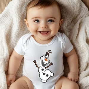 May include: A smiling baby wearing a white onesie with a graphic of Olaf from Frozen. The Olaf graphic is in black, brown, orange, and white. The baby is lying on a white blanket.