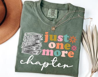 Puede incluir: Camiseta verde oliva con el texto "just one more chapter" y un gráfico de una pila de libros. El texto está en una mezcla de colores, incluyendo naranja, rosa y turquesa. Se ve un collar dorado.