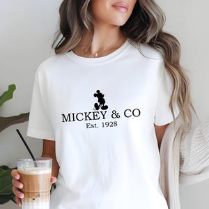 May include: White t-shirt featuring a black Mickey Mouse silhouette and the text "MICKEY & CO Est. 1928". The shirt is a classic crew neck style.