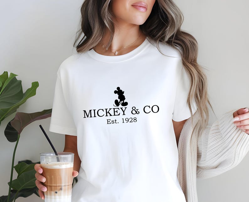 May include: White t-shirt featuring a black Mickey Mouse silhouette and the text "MICKEY & CO Est. 1928". The shirt is a classic crew neck style.