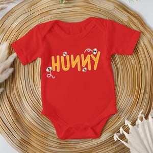 May include: A red baby onesie with the word "HUNNY" in yellow, with bee graphics. The onesie is short-sleeved and has snaps at the bottom. The background is a woven, circular mat.