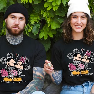Può includere: Due magliette nere abbinate con un design a cartoni animati di Topolino e Minnie. La grafica mostra i personaggi che si abbracciano e si baciano, con un cuore sopra. Una persona indossa un berretto nero, l'altra un berretto bianco.