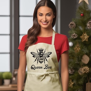 May include: Beige apron with a black graphic of a bee wearing a crown, and the text "Queen Bee". The apron is tied at the waist and has a neck strap. The person is wearing a red shirt.