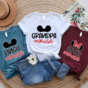 Può includere: Tre magliette grafiche con orecchie di Topolino e la scritta "Uncle Mouse", "Grandpa Mouse" e "Auntie Mouse". Le magliette sono blu, bianche e bordeaux. Un paio di jeans blu e un cappello di paglia sono anche nell'immagine.