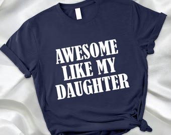 May include: A navy blue t-shirt with the white text "AWESOME LIKE MY DAUGHTER". The shirt is made of a soft material and has rolled-up sleeves. The shirt is knotted at the bottom.