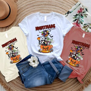 May include: Three t-shirts in white, cream, and coral, featuring a Halloween-themed graphic. The design includes characters from "The Nightmare Before Christmas" rising from a jack-o'-lantern cup, with the text "Nightmare on Main Street". Blue jeans and a straw hat are also in the image.