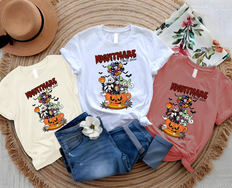 May include: Three t-shirts in white, cream, and coral, featuring a Halloween-themed graphic. The design includes characters from "The Nightmare Before Christmas" rising from a jack-o'-lantern cup, with the text "Nightmare on Main Street". Blue jeans and a straw hat are also in the image.