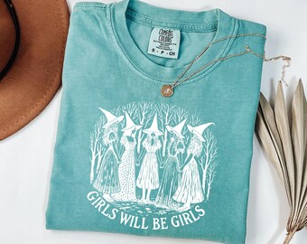 May include: A teal Comfort Colors t-shirt featuring a white graphic of five witches in dresses and witch hats, holding hands in a circle. The text "GIRLS WILL BE GIRLS" is below the graphic. A gold necklace and a brown hat are also visible.