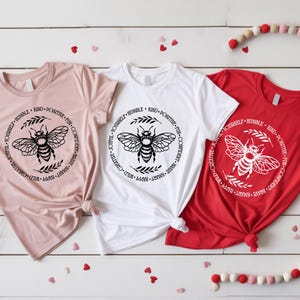 May include: Three t-shirts in pink, white, and red, each featuring a circular design with a bee illustration and the words "Kind, Positive, Fun, Confident, Brave, Smart, Happy, Wild, Creative, Joyful, Yourself, Humble." The shirts are knotted at the waist.