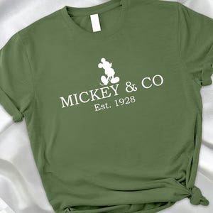 May include: Olive green t-shirt with the text "MICKEY & CO Est. 1928" and a white silhouette of Mickey Mouse. The shirt has rolled-up sleeves and a knotted side.