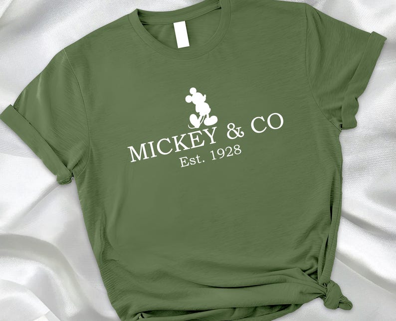May include: Olive green t-shirt with the text "MICKEY & CO Est. 1928" and a white silhouette of Mickey Mouse. The shirt has rolled-up sleeves and a knotted side.