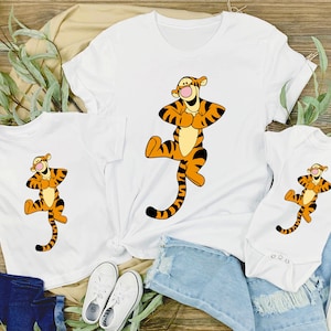May include: White t-shirts and a onesie featuring a cartoon Tigger design. The Tigger character is orange with black stripes and is in a playful pose. The shirts are displayed on a wooden surface with greenery.