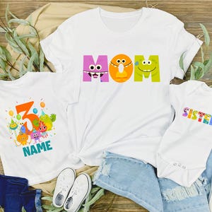 May include: White t-shirts and a onesie with colorful cartoon monster designs. One shirt says "MOM" in large letters, another has a number 5 with "NAME" below, and the onesie says "SISTER". The shirts are on a wooden surface with denim shorts and white sneakers.