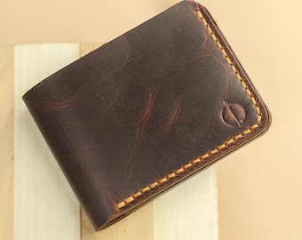 May include: A dark brown leather bifold wallet featuring a smooth, distressed texture and thick, contrasting yellow stitching along the edges. A small, circular embossed logo is visible on the bottom right corner of the front panel.